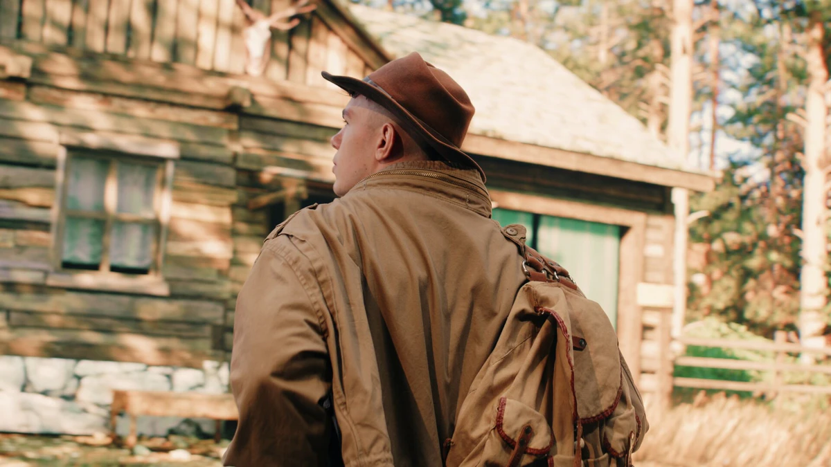 Shot: a man in a hat in front of a wooden house